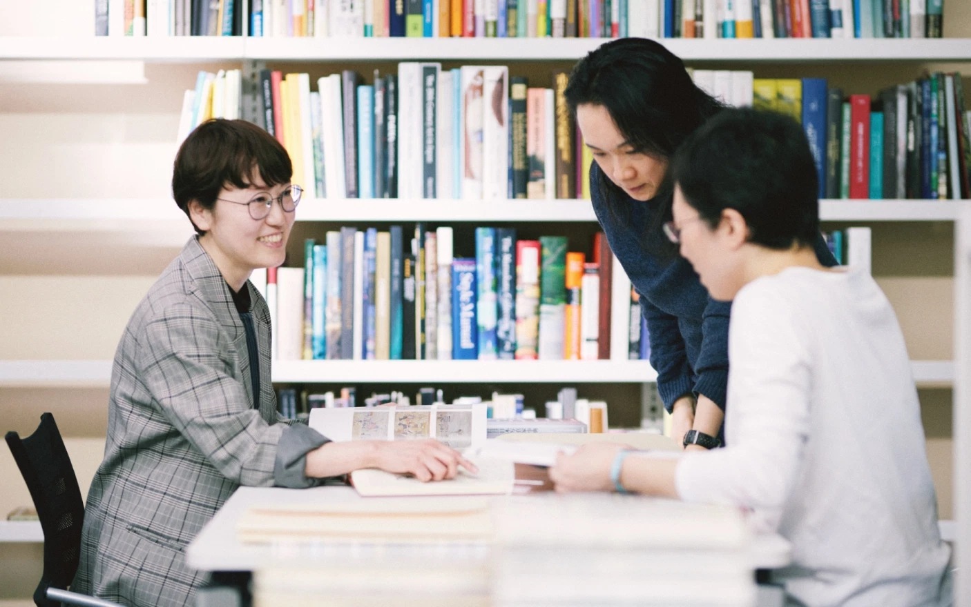 Three people looking at books spread out on a desk and having a discussion while pointing with their fingers.